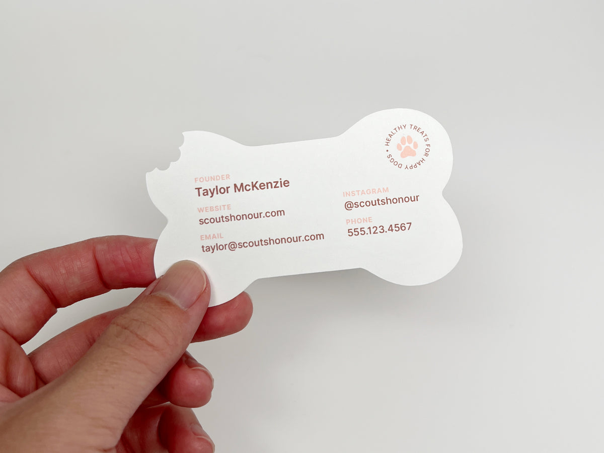 A hand holds the back view of a bone-shaped business card for Scout Honour, featuring a white background with coral text and logo, against a light gray studio backdrop.