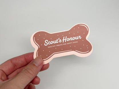 Hand holding a brown, bone-shaped dog bakery business card reading “Scout’s Honour—Healthy Treats for Happy Dogs.”