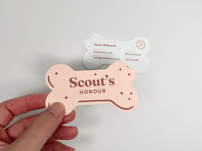 A hand holds a bone-shaped pink card reading 'Scout's Honour' in front of a white bone-shaped card on a plain light background.