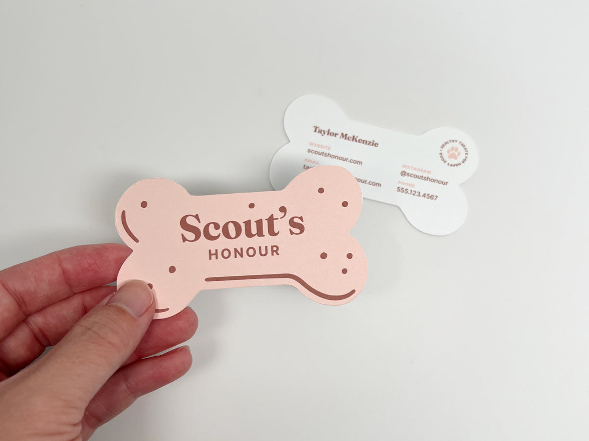 Hand holding a pink, bone-shaped dog bakery business card reading “Scout’s Honour,” with the white contact-information side displayed behind it.