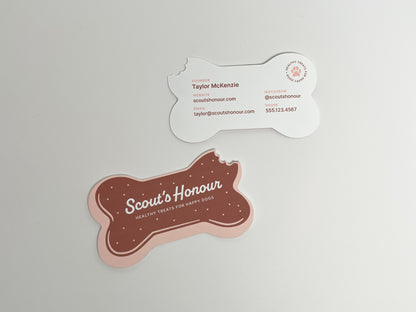 Front and back of a bitten, bone-shaped dog bakery business card featuring a brown treat design and editable contact information.
