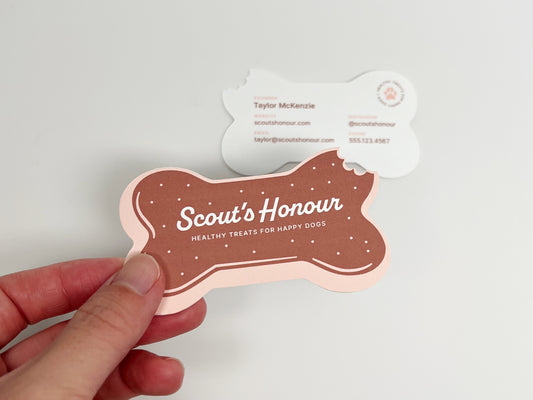 A hand holds a bone-shaped printed card in front of a white background. The front card is dusty rose pink with a brown center and white text reading 'Scout's Honour Healthy Treats for Happy Dogs.'