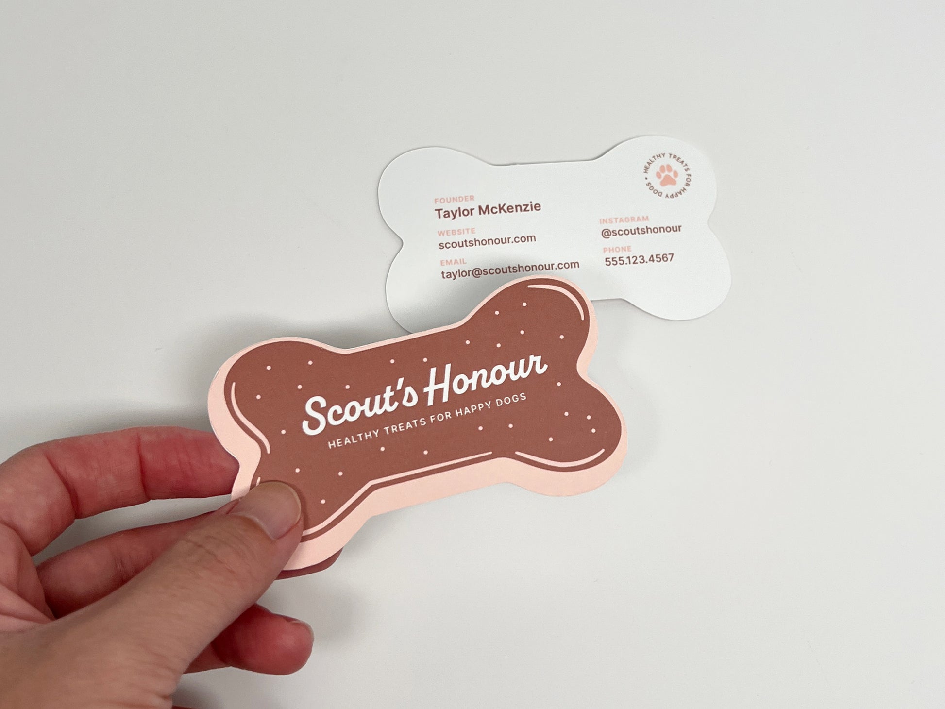 A hand holds a bone-shaped pink and brown card in front of a white bone-shaped card on a plain light background. The front card reads 'Scout's Honour' with smaller text 'Healthy Treats for Happy Dogs' and includes the Scout's Honour logo.