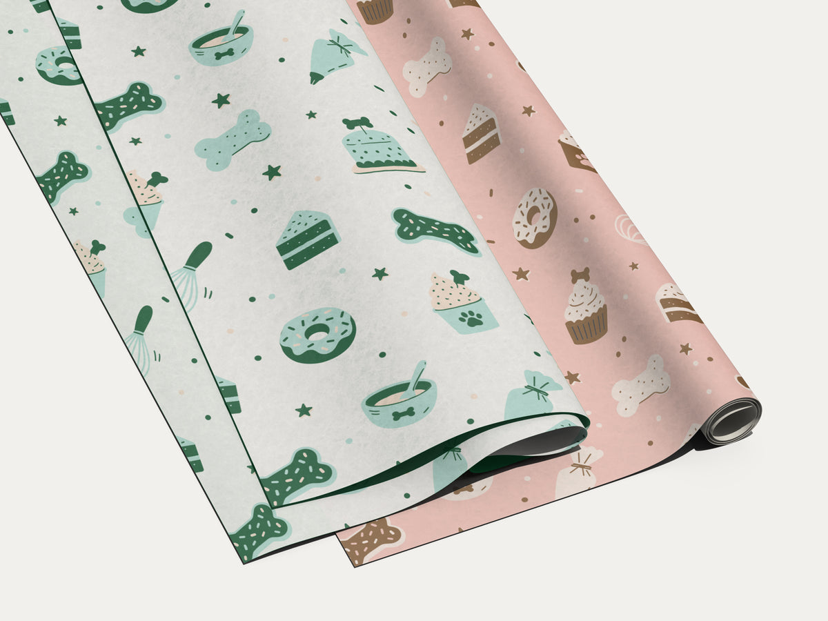 Seamless dog bakery pattern in white and pink colorways, shown side by side as folded white sheets and a partly unrolled pink wrapping paper roll. The design features bone cookies, paw-print cupcakes, cake slices, donuts, treat bowls, piping bags, whisks, and stars.