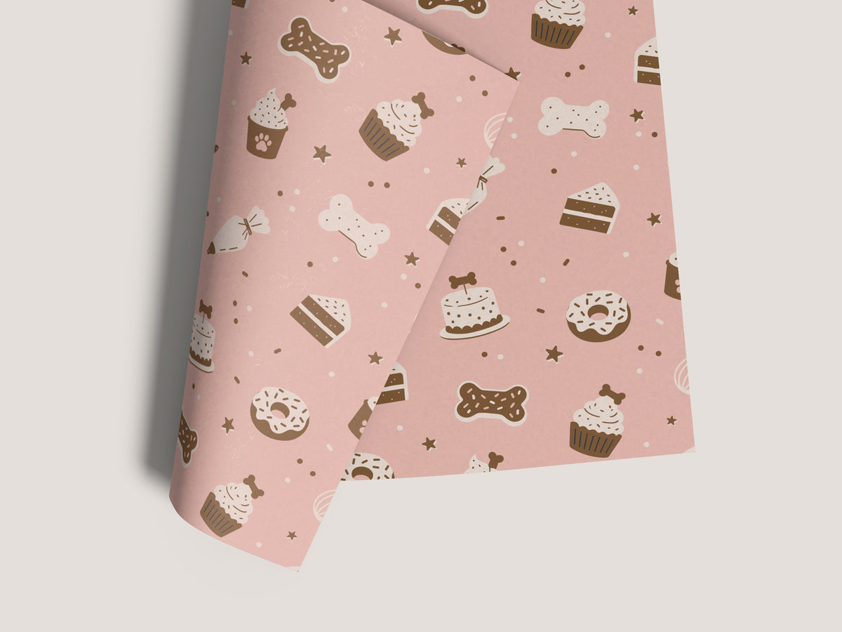 Seamless dog bakery pattern in the pink colorway, shown on a sheet of paper with a curled corner on a light gray background. The design features bone-shaped cookies, bone-topped cakes and cupcakes, paw-print cupcakes, cake slices, donuts, piping bags, and stars in brown and cream.