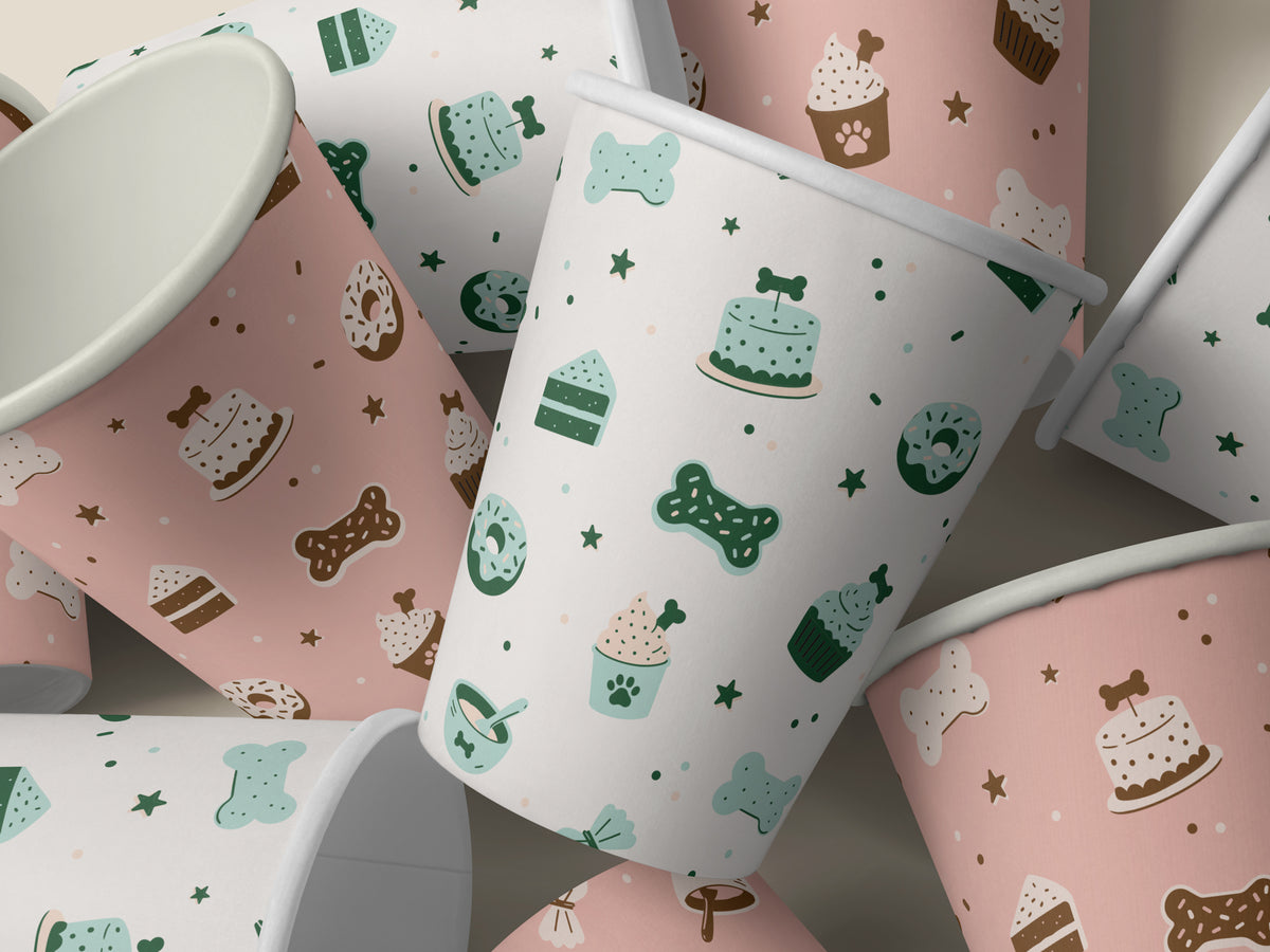Seamless dog bakery pattern shown on pink and white paper cups. The design features bone-shaped cookies, bone-topped cakes, paw-print cupcakes, donuts, and stars in mint, green, and brown.