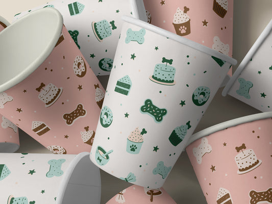 Seamless dog bakery pattern shown on pink and white paper cups. The design features bone-shaped cookies, bone-topped cakes, paw-print cupcakes, donuts, and stars in mint, green, and brown.
