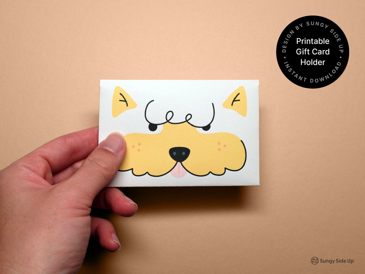 Close-up of a hand holding the finished printable dog gift card holder against a soft peach background. The card features an illustrated yellow dog face with expressive eyes and a black nose. A round black label in the top right corner reads Printable Gift Card Holder and mentions Instant Download and Designed by Sungy Side Up. This image shows the final product and highlights the charming handmade design.