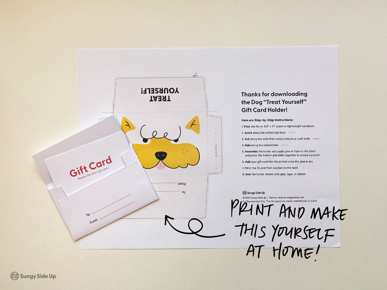 Flat lay of the printable dog gift card holder template placed on a light surface. A completed card holder is shown with a sample gift card partially inserted. A playful handwritten-style message on the photo says Print and make this yourself at home. The template includes a dog face design and step-by-step instructions. This image highlights the ease and fun of assembling the digital download yourself.