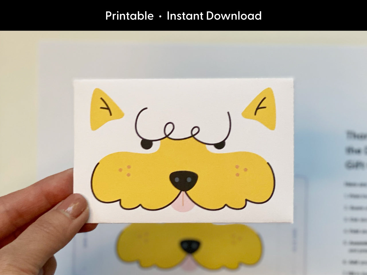 Hand holding the finished printable dog gift card holder featuring a yellow cartoon dog face with curly eyebrows and a pink tongue. The background is softly blurred and includes the original template to show how it looks before being assembled. At the top of the image, white text on black reads Printable Instant Download to emphasize the digital nature of the product. Great for showing the final result.