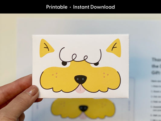 Hand holding the finished printable dog gift card holder featuring a yellow cartoon dog face with curly eyebrows and a pink tongue. The background is softly blurred and includes the original template to show how it looks before being assembled. At the top of the image, white text on black reads Printable Instant Download to emphasize the digital nature of the product. Great for showing the final result.