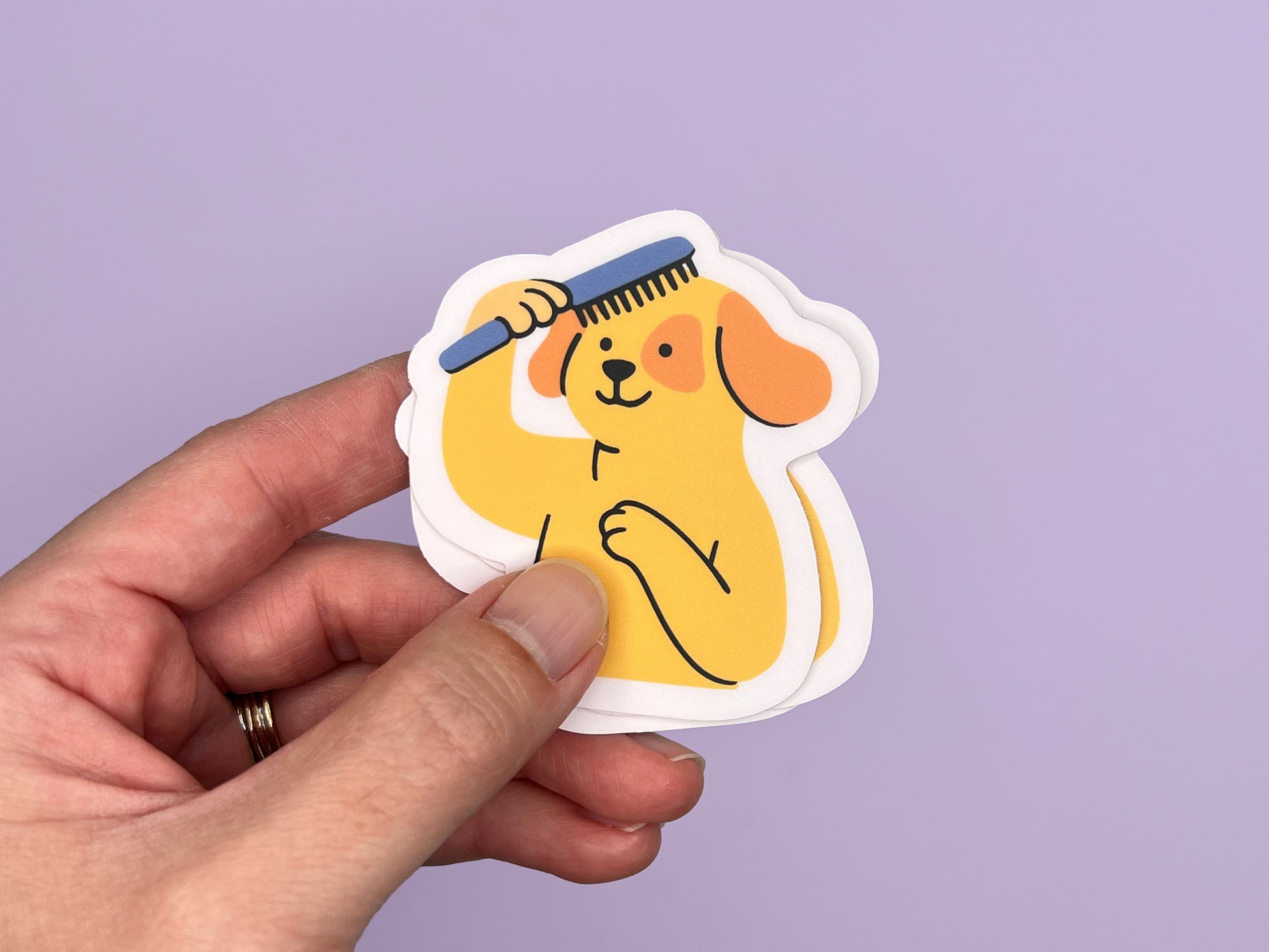 Hand holding a small stack of matte vinyl die cut stickers featuring an illustrated smiling yellow dog brushing its own hair with a blue comb. The dog has floppy orange ears and one paw on its chest. Each sticker has a soft pink border and is shown against a light purple background, highlighting the sticker’s thickness and compact size.