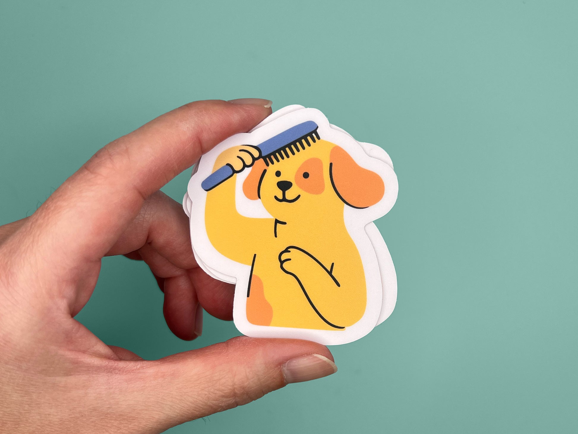 Hand holding a small matte vinyl die cut sticker featuring an illustrated smiling yellow dog brushing its own hair with a blue comb. The dog has floppy orange ears and stands upright with one paw on its chest. The sticker has a soft pink border and is shown against a teal background, highlighting its compact size and playful design.