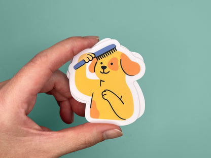 Hand holding a small matte vinyl die cut sticker featuring an illustrated smiling yellow dog brushing its own hair with a blue comb. The dog has floppy orange ears and stands upright with one paw on its chest. The sticker has a soft pink border and is shown against a teal background, highlighting its compact size and playful design.