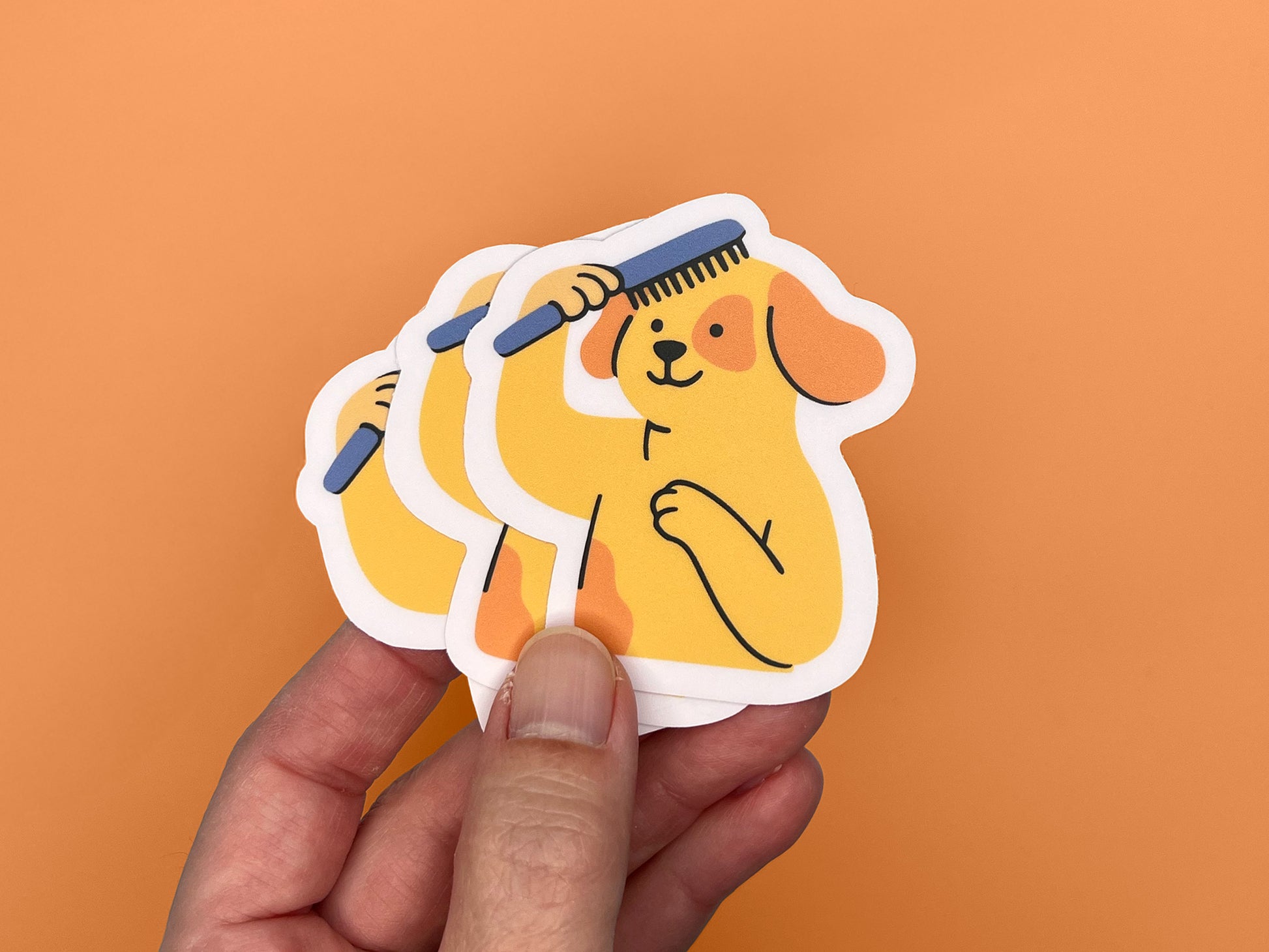Hand holding three matte vinyl die cut stickers featuring an illustrated smiling yellow dog brushing its own hair with a blue comb. The dog has floppy orange ears and one paw on its chest. Each sticker has a soft pink border and is shown against an orange background, highlighting the layered stickers and compact size.