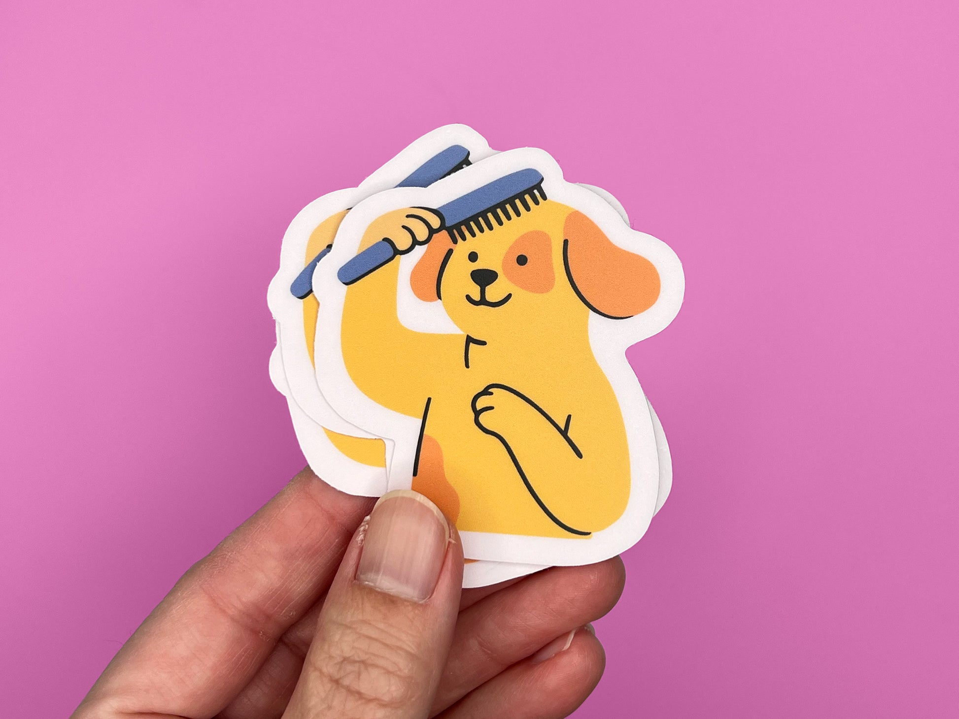 Hand holding a small stack of matte vinyl die cut stickers featuring an illustrated smiling yellow dog brushing its own hair with a blue comb. The dog has floppy orange ears and one paw on its chest. Each sticker has a soft pink border and is shown against a bright pink background, highlighting the layered stickers and compact size.