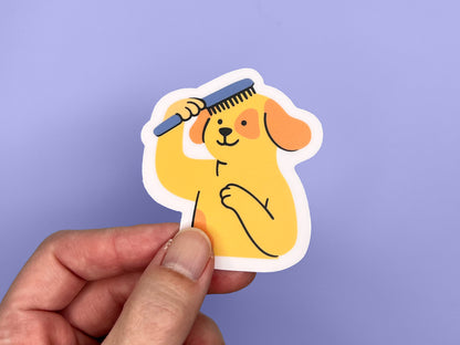 Hand holding a single matte vinyl die cut sticker featuring an illustrated smiling yellow dog brushing its own hair with a blue comb. The dog has floppy orange ears and one paw on its chest. The sticker has a soft pink border and is shown against a light purple background, highlighting the sticker’s size and playful design.