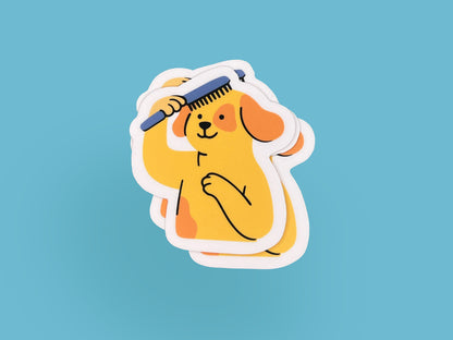 Small stack of matte vinyl die cut stickers featuring an illustrated smiling yellow dog brushing its own hair with a blue comb. The dog has floppy orange ears and one paw on its chest. Each sticker has a soft pink border and is shown on a light blue background, highlighting the layered stack and compact size.