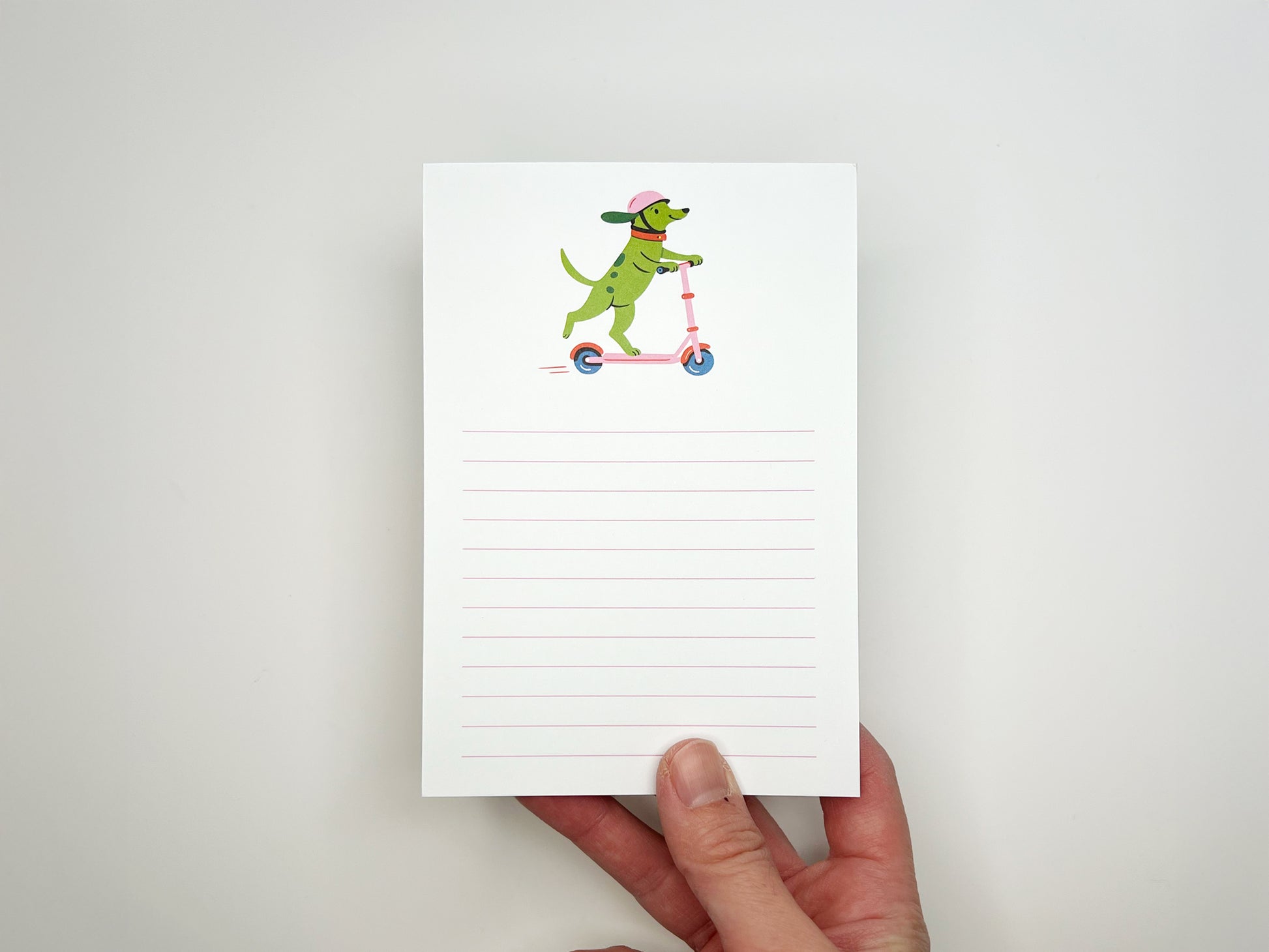 Hand holding a 4 × 6 inch lined notepad featuring an illustrated green dog wearing a pink helmet and orange collar while riding a pink scooter. The memo pad has soft pink horizontal lines for writing and a white background, photographed against a light neutral surface.