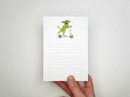 Hand holding a 4 × 6 inch lined notepad featuring an illustrated green dog wearing a pink helmet and orange collar while riding a pink scooter. The memo pad has soft pink horizontal lines for writing and a white background, photographed against a light neutral surface.