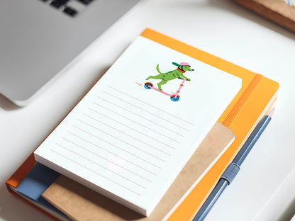 Small lined notepad with an illustration of a green dog wearing a pink helmet and riding a pink scooter, resting on top of stacked notebooks. A pen lies beside the notebooks, and part of a laptop is visible in the corner on a white desk.