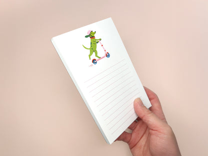 Hand holding a thick 4 × 6 inch lined notepad at an angle, showing an illustrated green dog wearing a pink helmet and orange collar while riding a pink scooter. The memo pad features soft pink horizontal lines on white paper and is photographed against a peach-colored background.