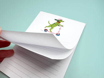 A hand flips through a small lined notepad. The top page shows an illustration of a green dog wearing a pink helmet and riding a pink scooter. Several blank lined pages are visible beneath, photographed against a light teal background.