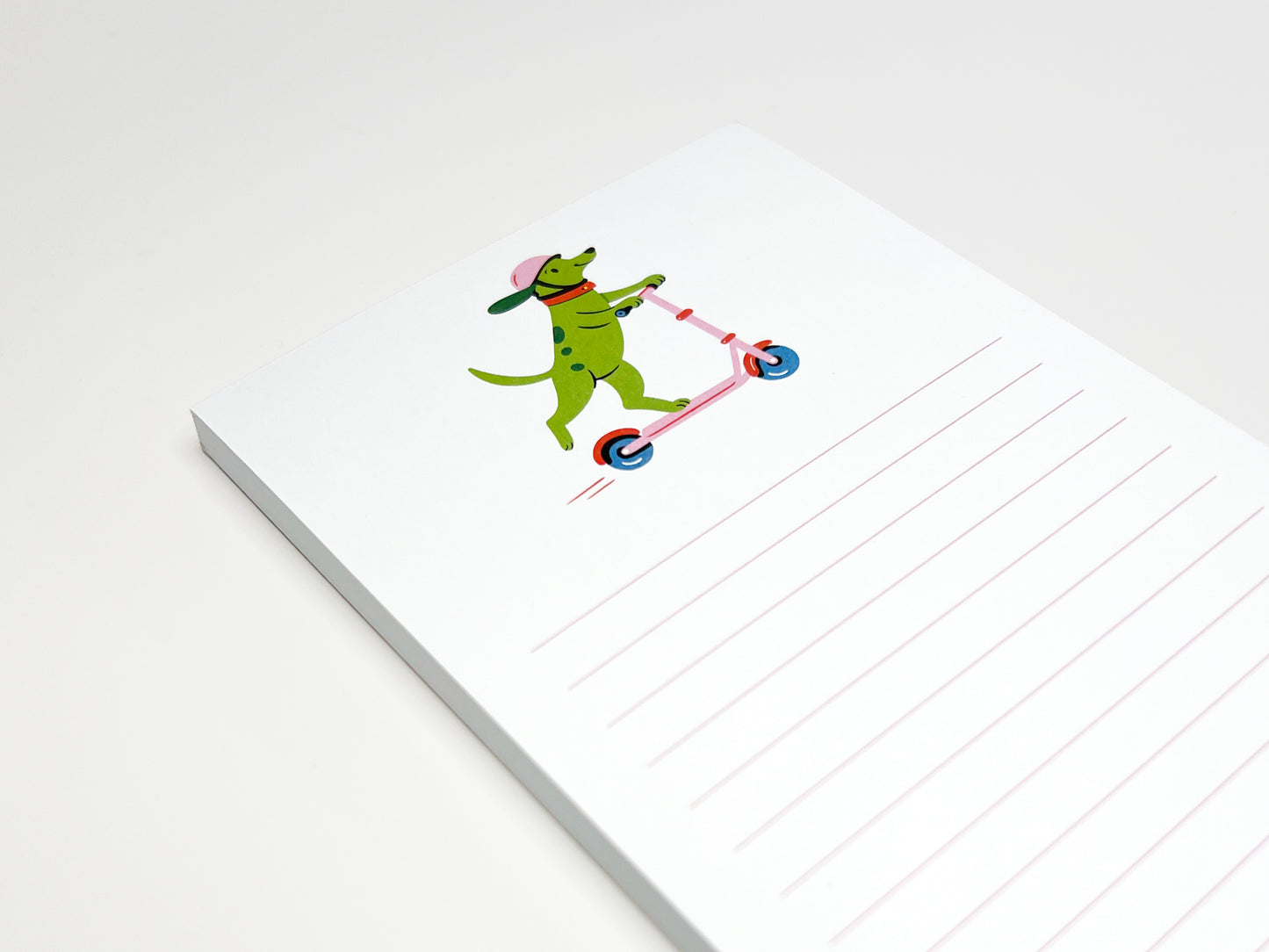 Close-up of a small lined notepad on a white surface. The top page shows an illustration of a green dog wearing a pink helmet and riding a pink scooter. Pale pink horizontal lines fill the lower half of the page.