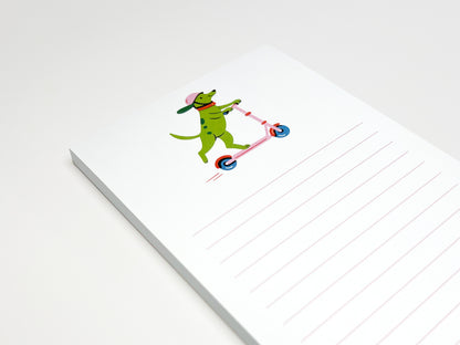 Close-up of a small lined notepad on a white surface. The top page shows an illustration of a green dog wearing a pink helmet and riding a pink scooter. Pale pink horizontal lines fill the lower half of the page.