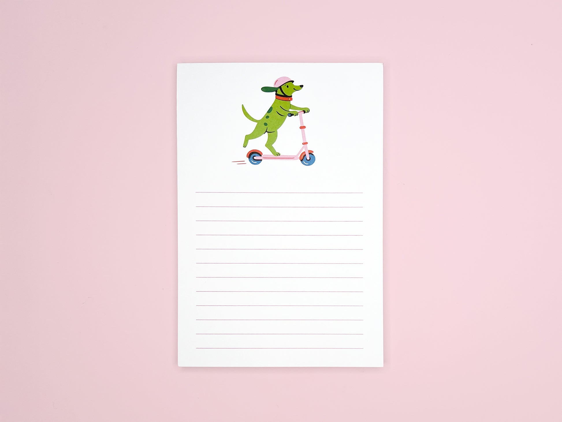 Small lined notepad centered on a light pink background. The top of the page shows an illustration of a green dog wearing a pink helmet and riding a pink scooter. Pale pink horizontal lines fill the lower portion of the page.