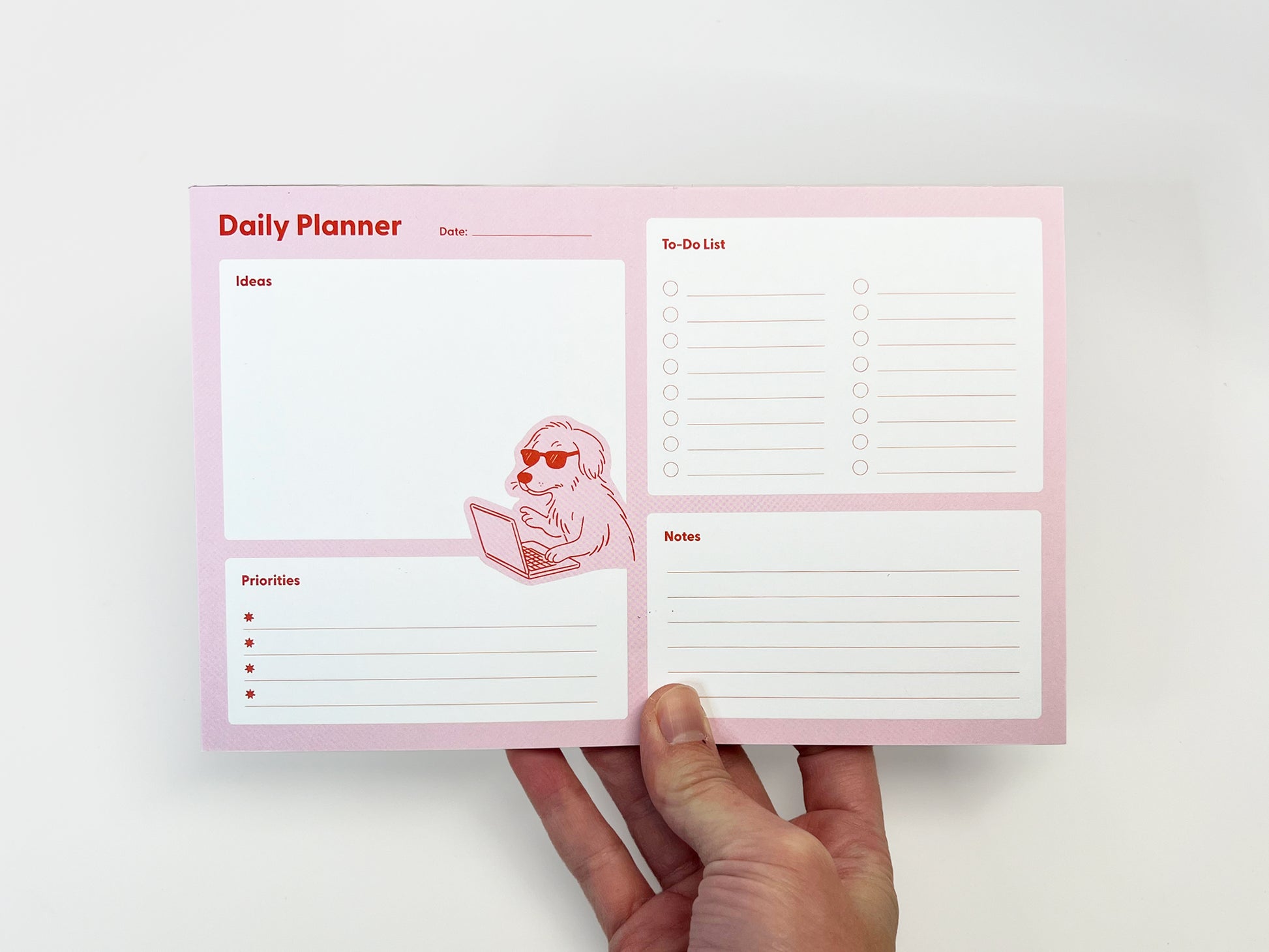 Front view of a pink daily planner notepad held in one hand against a white background, featuring labeled sections for Ideas, Priorities, To-Do List, and Notes. A small illustration of a dog wearing sunglasses and typing on a laptop appears near the center. The planner has a clean, structured layout designed for daily planning and note-taking.