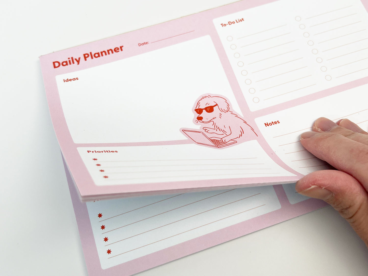 Close-up of a pink daily planner notepad with multiple tear-off sheets, showing a page being lifted to reveal the stack underneath. The layout includes sections labeled Ideas, Priorities, To-Do List, and Notes. A playful illustration of a dog wearing sunglasses and typing on a laptop appears near the center, highlighting the planner’s fun, functional design.
