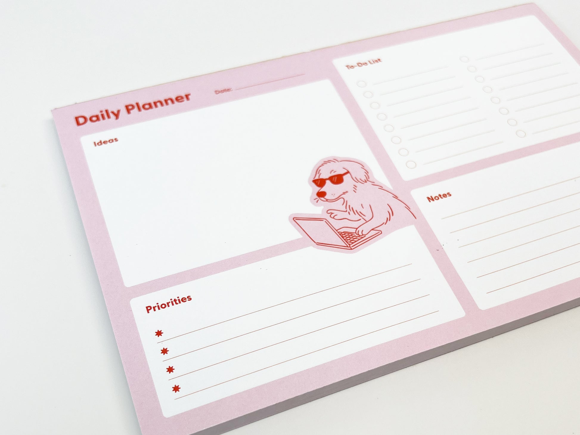 Angled close-up of a pink daily planner notepad on a white surface, featuring sections labeled Ideas, Priorities, To-Do List, and Notes. A playful illustration of a dog wearing sunglasses and typing on a laptop appears near the center. The planner’s structured layout and soft pink color palette are clearly visible.