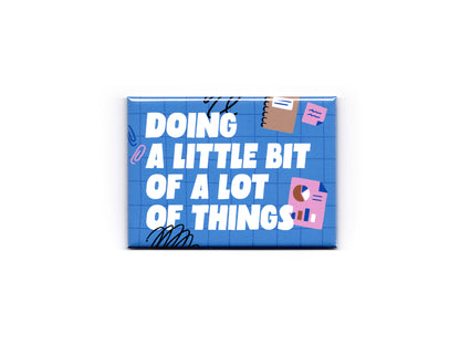 Blue rectangular magnet with bold white text reading “Doing a Little Bit of a Lot of Things” on a blue grid background. The design includes playful office-themed illustrations such as sticky notes, charts, paper clips, and black scribble accents against a light gray background.