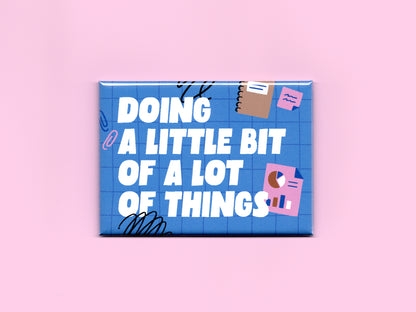 Blue rectangular magnet on a pink background with bold white text reading “Doing a Little Bit of a Lot of Things.” The design features a blue grid pattern with playful office-themed illustrations including sticky notes, charts, paper clips, and black scribble accents.