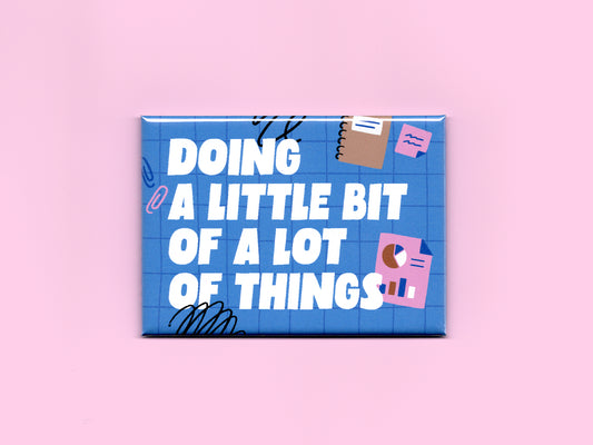 Blue rectangular magnet on a pink background with bold white text reading “Doing a Little Bit of a Lot of Things.” The design features a blue grid pattern with playful office-themed illustrations including sticky notes, charts, paper clips, and black scribble accents.