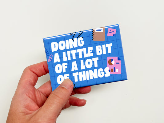 Hand holding a blue rectangular magnet with bold white text reading “Doing a Little Bit of a Lot of Things.” The magnet features a blue grid background with playful office-themed illustrations including charts, sticky notes, paper clips, and scribble accents against a plain white background.