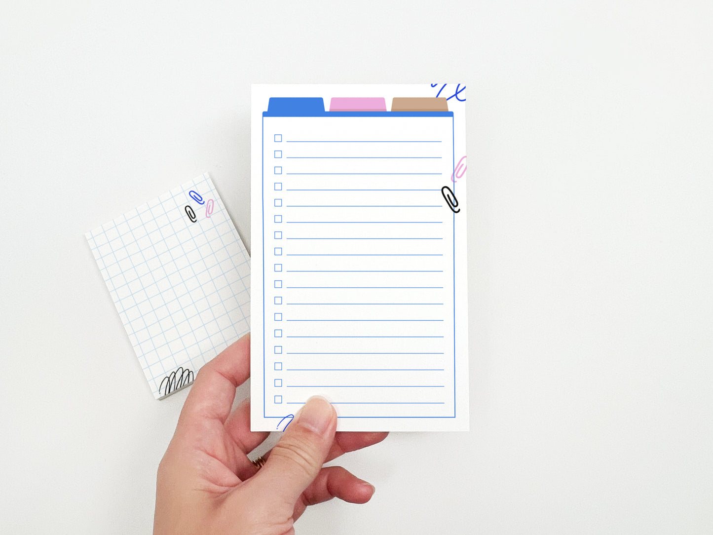 Hand holding a lined checklist notepad with colorful office-themed illustrations, with a matching mini grid notepad in the background.