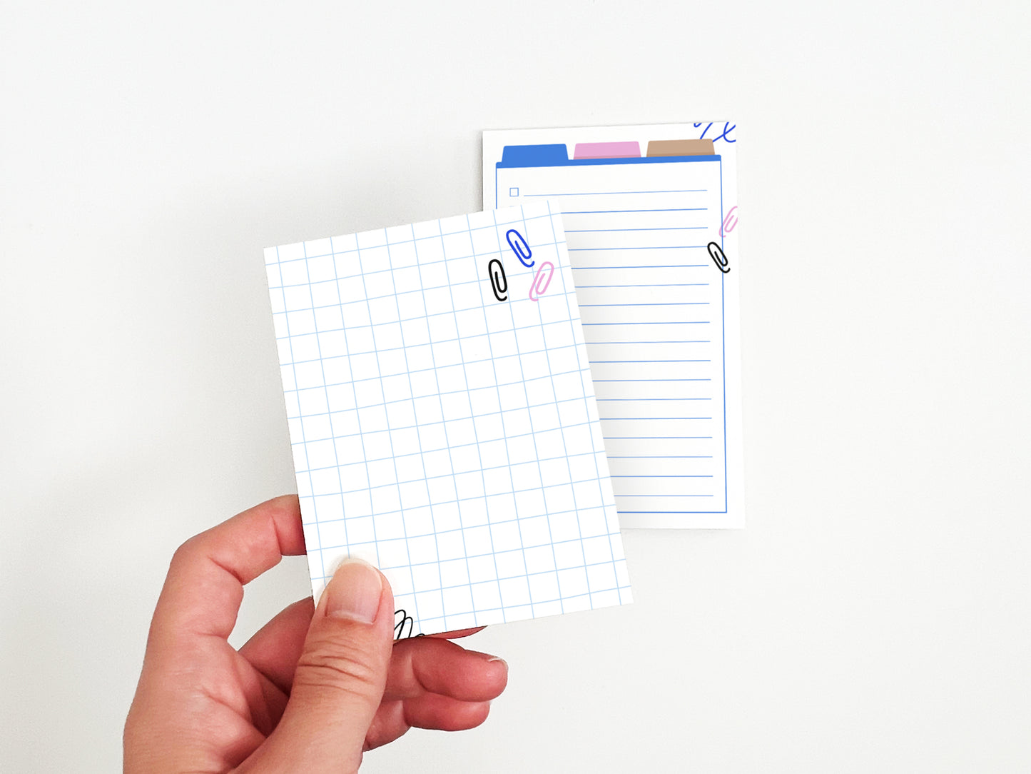 Hand holding a small grid-pattern notepad in front of a larger lined notepad with colorful office-themed illustrations on a white background.