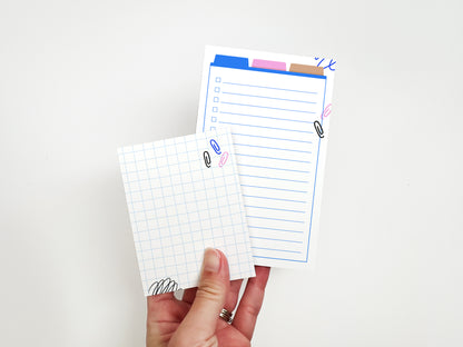 Hand holding a mini grid notepad and a lined checklist notepad with colorful office-themed illustrations on a white background.