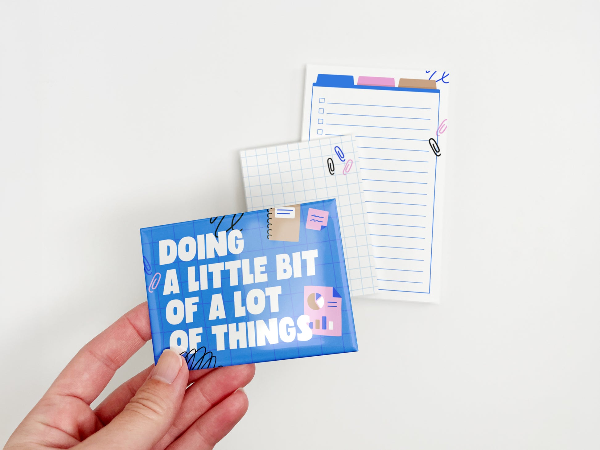 Hand holding a blue magnet that reads "Doing a Little Bit of a Lot of Things" with matching grid and lined notepads displayed in the background.