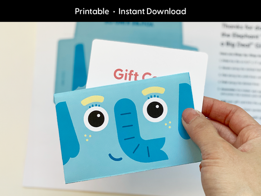 A hand holds a completed elephant-themed printable gift card holder with a visible gift card tucked inside. The cute blue elephant face features big eyes, yellow eyebrows, and a friendly smile. The background includes printed instructions slightly out of focus. This image showcases the finished product, perfect for birthdays, teacher appreciation, or baby showers. Ideal for promoting a fun and easy instant download.