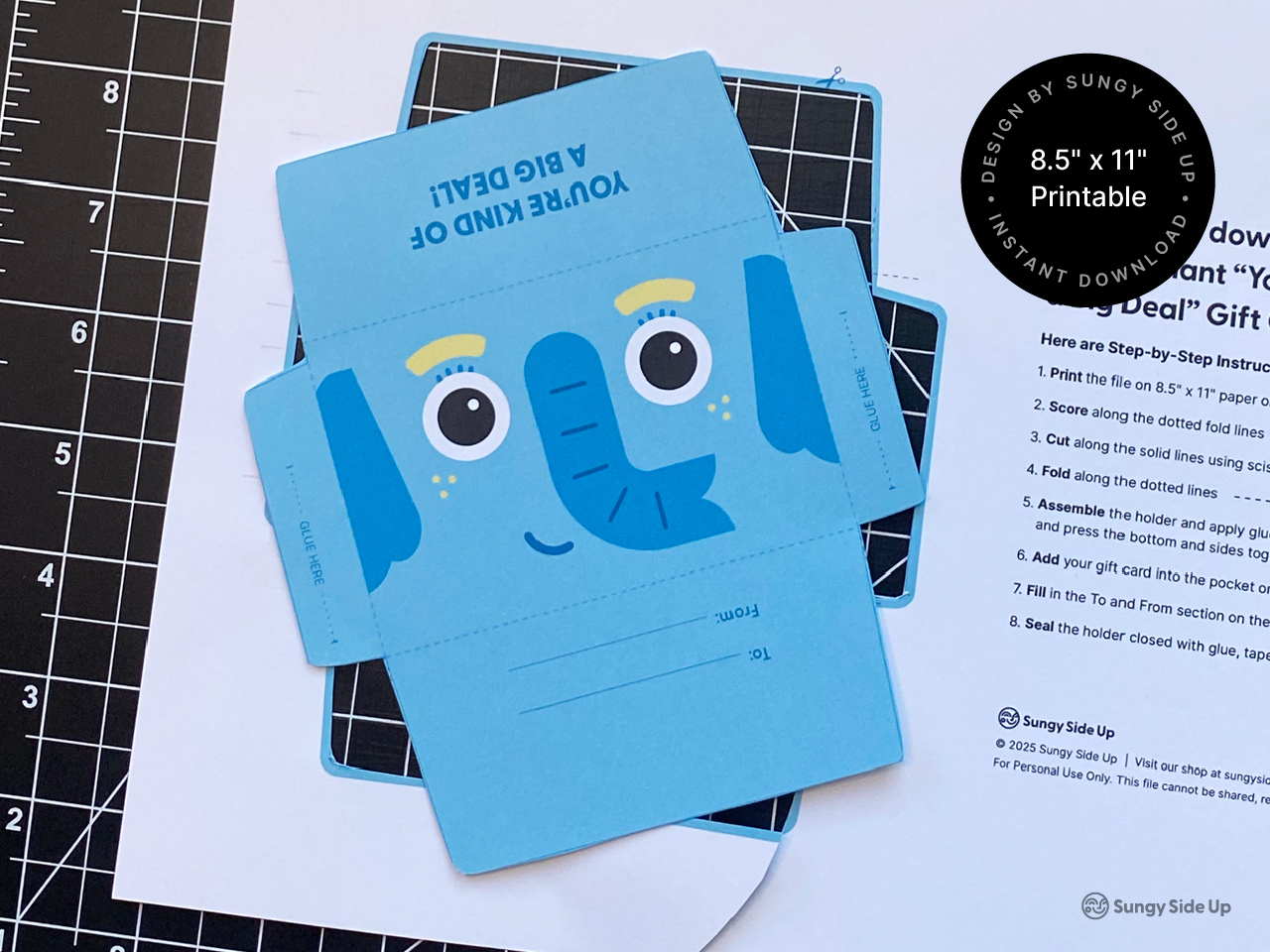 Flat lay image shows a printable elephant gift card holder in progress on a cutting mat. The design features a cheerful blue elephant with text that reads “You’re Kind of a Big Deal!” Instructions are printed on a white sheet beside it. A black circular label notes it’s an 8.5" x 11" printable for instant download. This setup highlights the DIY aspect, appealing to crafters and gift-givers looking for a creative, printable option.
