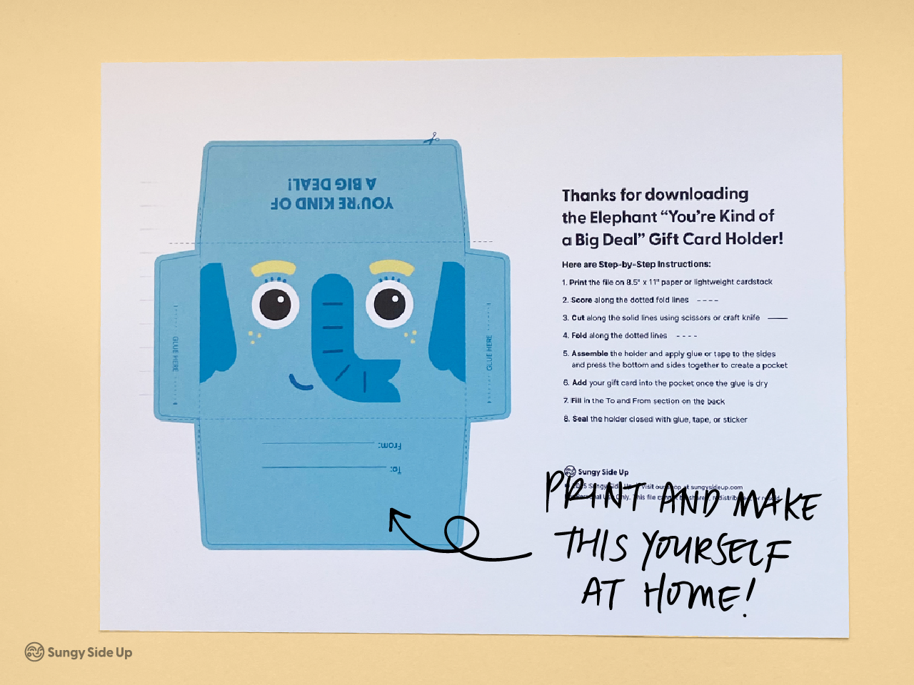 Flat lay of a printable elephant gift card holder on a yellow background. The design features a blue cartoon elephant with big eyes and “You’re Kind of a Big Deal” text. A step-by-step instruction sheet is visible next to the design. Handwritten text on the image says “Print and make this yourself at home!” This image highlights the DIY aspect of the product, ideal for buyers seeking fun and easy printable gifts.