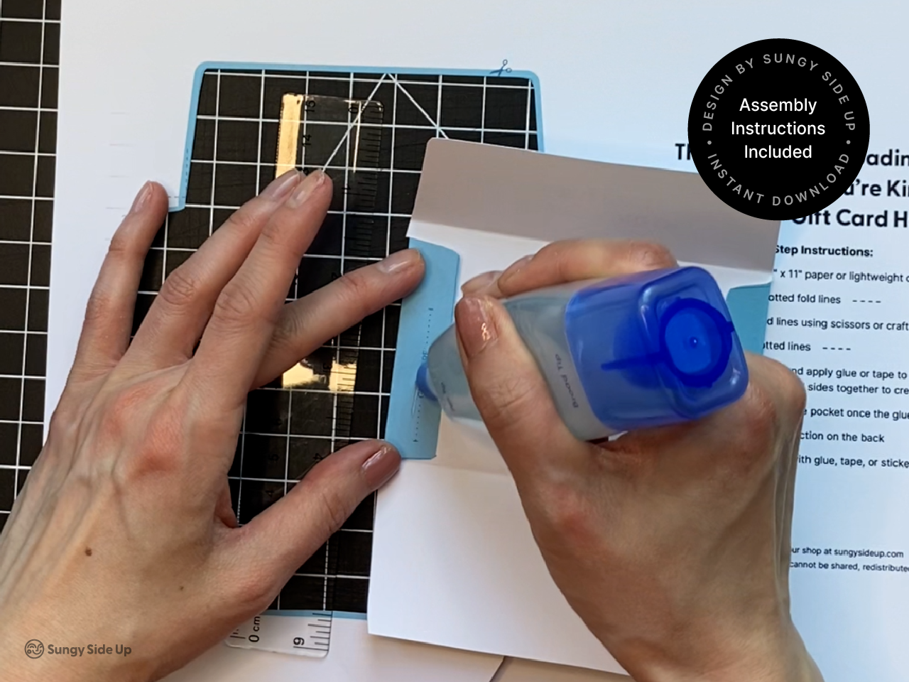 Close-up of hands applying glue to the edge of a printable elephant gift card holder template. The blue elephant design is placed on a cutting mat with a ruler and printed instructions in the background. A label in the corner reads “Assembly Instructions Included.” This photo showcases the hands-on DIY crafting process for customers looking to create their own fun and easy gift card holders at home.