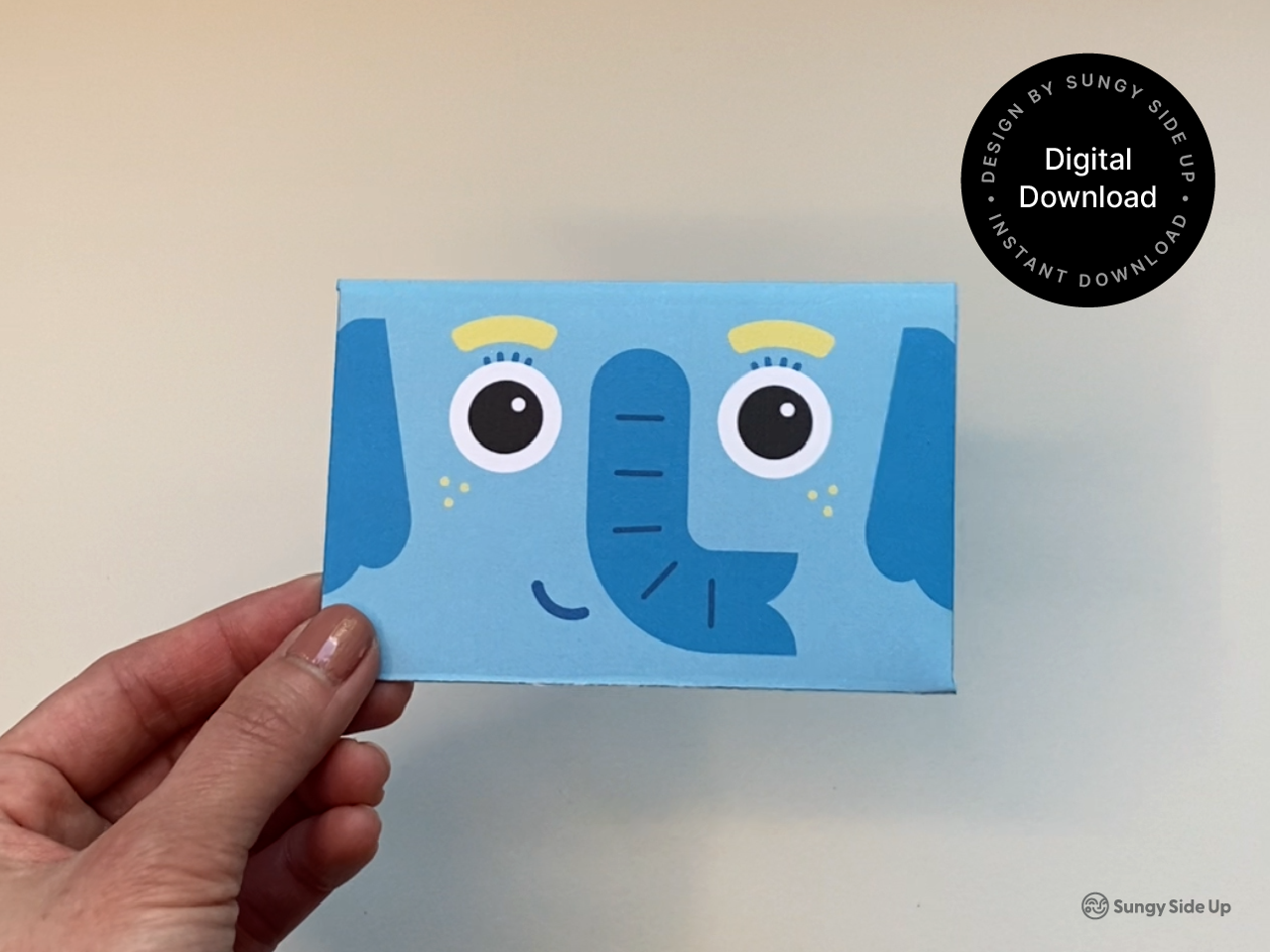 A hand holds the completed elephant gift card holder against a neutral background. The design features a bright blue elephant face with a friendly smile and large cartoon eyes. A circular badge reads “Digital Download – Instant Download – Design by Sungy Side Up.” This image highlights the finished product, perfect for Etsy shoppers searching for unique printable gift card holders and DIY gift ideas.