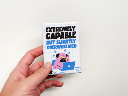 Hand holding a rectangular magnet with bold black and blue text reading “Extremely Capable But Slightly Overwhelmed.” The magnet features an illustration of a stressed pink pug wearing glasses while working on a blue laptop with scattered papers and a coffee mug on a white grid background.