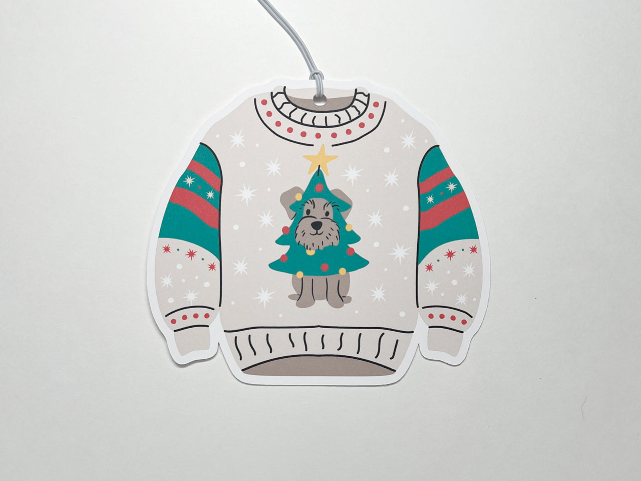 Christmas-themed sweater gift tag. The sweater is designed in light beige with festive decorations. On the front, there is a playful illustration of a small gray schnauzer dog dressed as a Christmas tree, complete with a yellow star on top. The sweater's sleeves feature red and green stripes with white snowflake patterns, matching the overall holiday aesthetic. This cute and festive design would be perfect for holiday gift-giving.