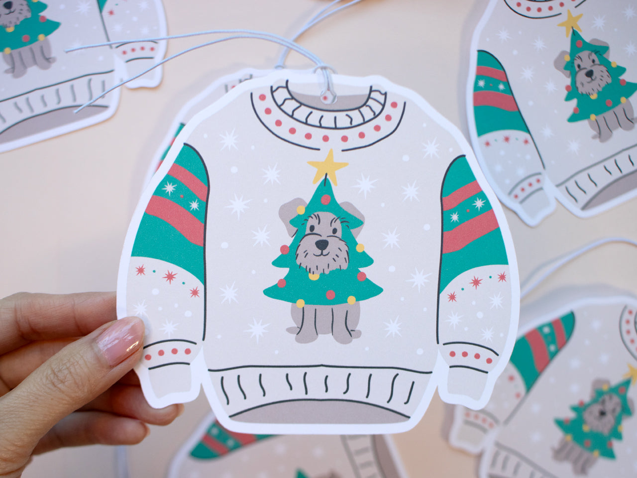 Hand holding a festive Christmas sweater-shaped gift tag featuring a playful design. The sweater is beige with green, red, and white holiday patterns on the sleeves. The main feature is a cute schnauzer dog dressed as a Christmas tree, complete with colorful ornaments and a yellow star on top. In the background, additional gift tags are scattered, showcasing the same design. The tag includes white snowflakes and is attached with a string for easy use on gifts.