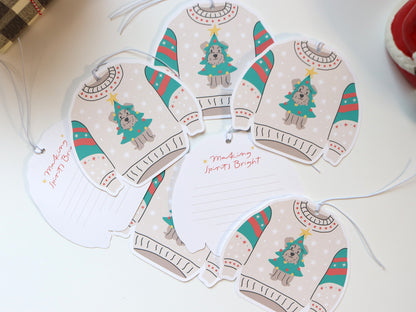 Several Christmas sweater-shaped gift tags spread across a surface. Each tag features a schnauzer dog dressed as a Christmas tree with ornaments and a star on top, set against a light beige sweater adorned with festive red, green, and white patterns. Some tags are flipped to the back, revealing lined spaces for personalized messages. One message reads “Making Spirits Bright” in red script. The tags are attached with white strings, ready to be used for holiday gift wrapping.